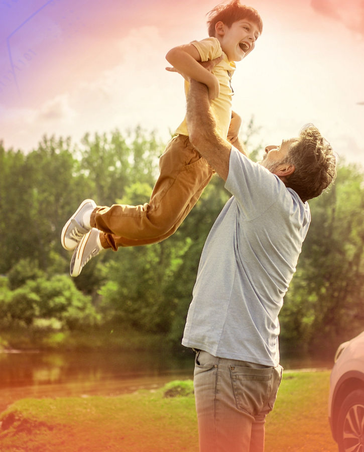 A person lifts a child joyfully in a scenic outdoor setting with lush greenery, a wind turbine in the background, and a car parked nearby, symbolizing family moments and sustainable living.