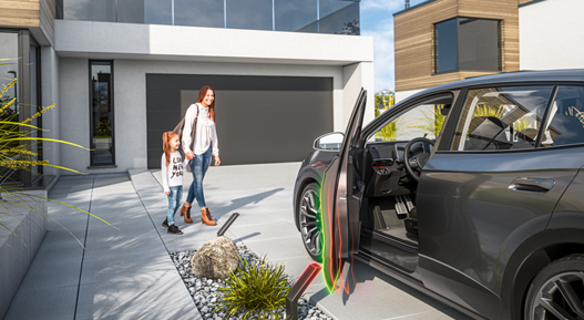 Modern car parked in front of a contemporary house with the driver’s door open, highlighted by green and red visual effects representing power door technology. Decorative landscaping and clean driveway complete the scene.